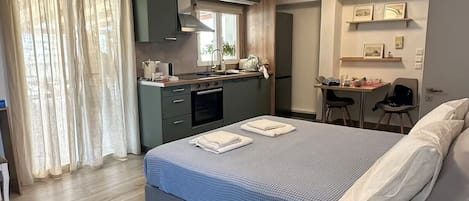 A modern hotel room with a large bed, a desk, a kitchenette, and a seating area.