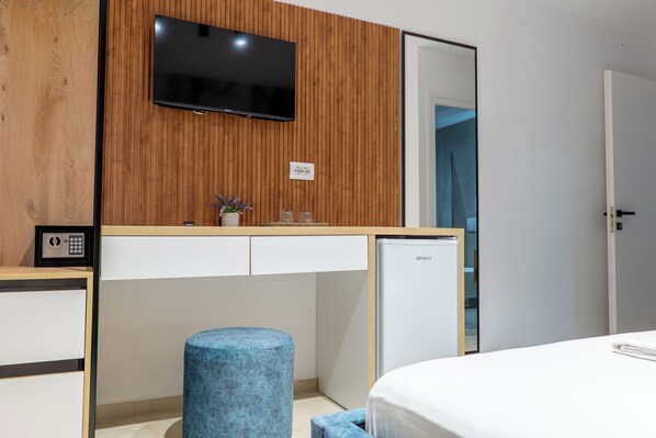 A modern hotel room with a wooden panel wall, a flat-screen TV, a minibar, a small blue stool, a bed, and a door leading to a bathroom. - Vila Naço Ksamil (Ksamil)