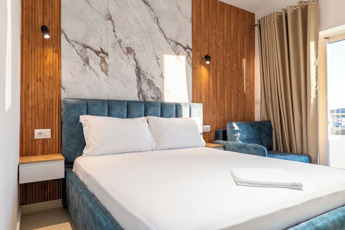 Habitación de hotel con pared de mármol, paneles de madera, una cama grande con ropa de cama blanca, cabecera azul, mesita de noche y un sillón azul.