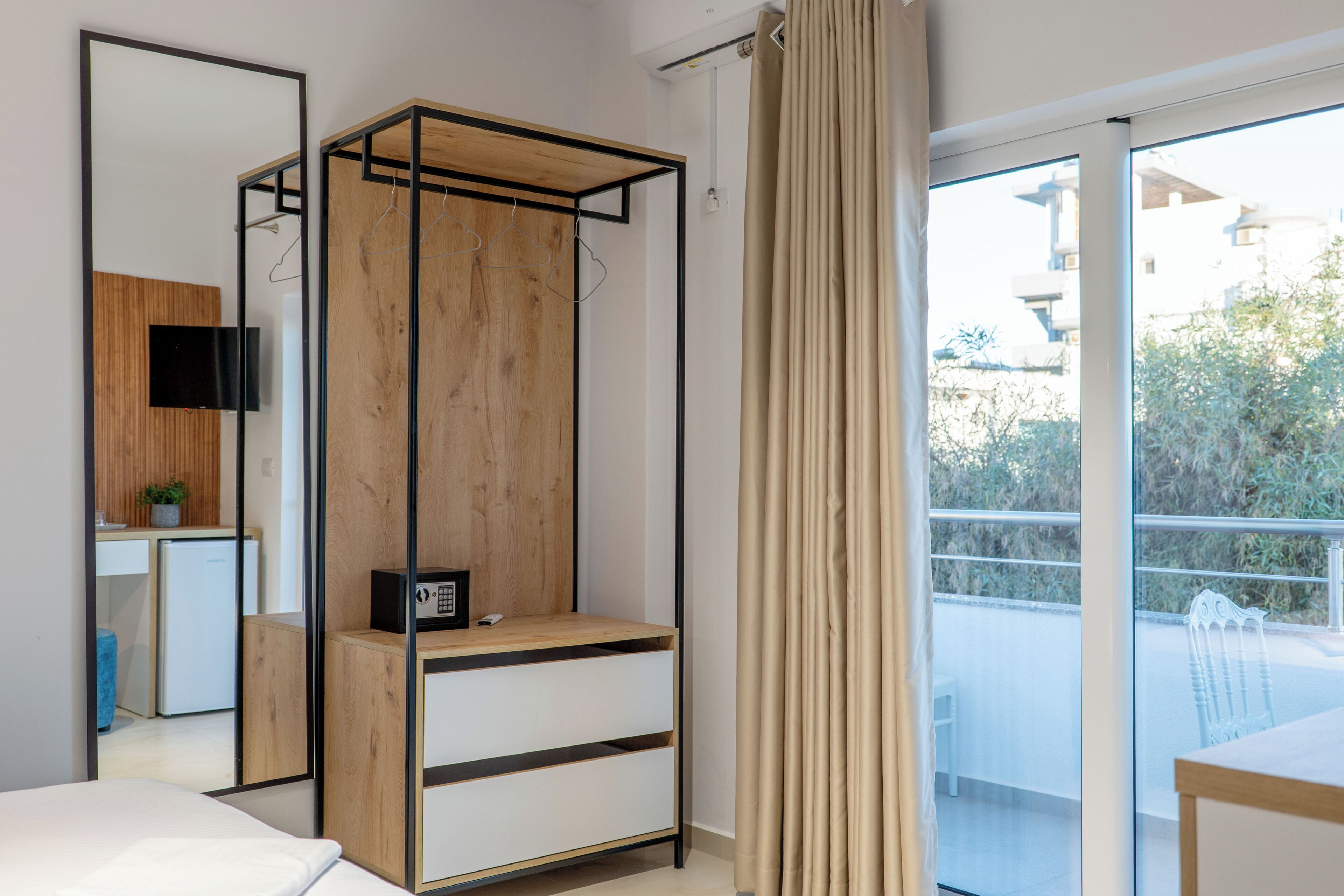A modern bedroom with a bed, a wooden wardrobe, a desk with a chair, a TV, and a balcony with a view of greenery.