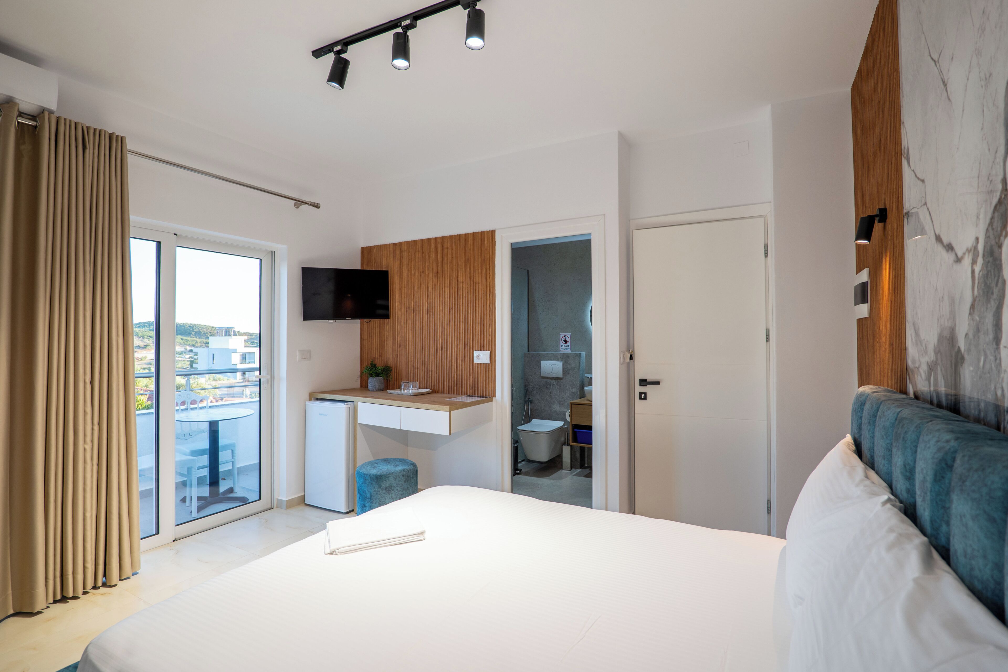 A modern hotel room with a large bed, a balcony with curtains, a small kitchenette, a bathroom, and a TV mounted on the wall.