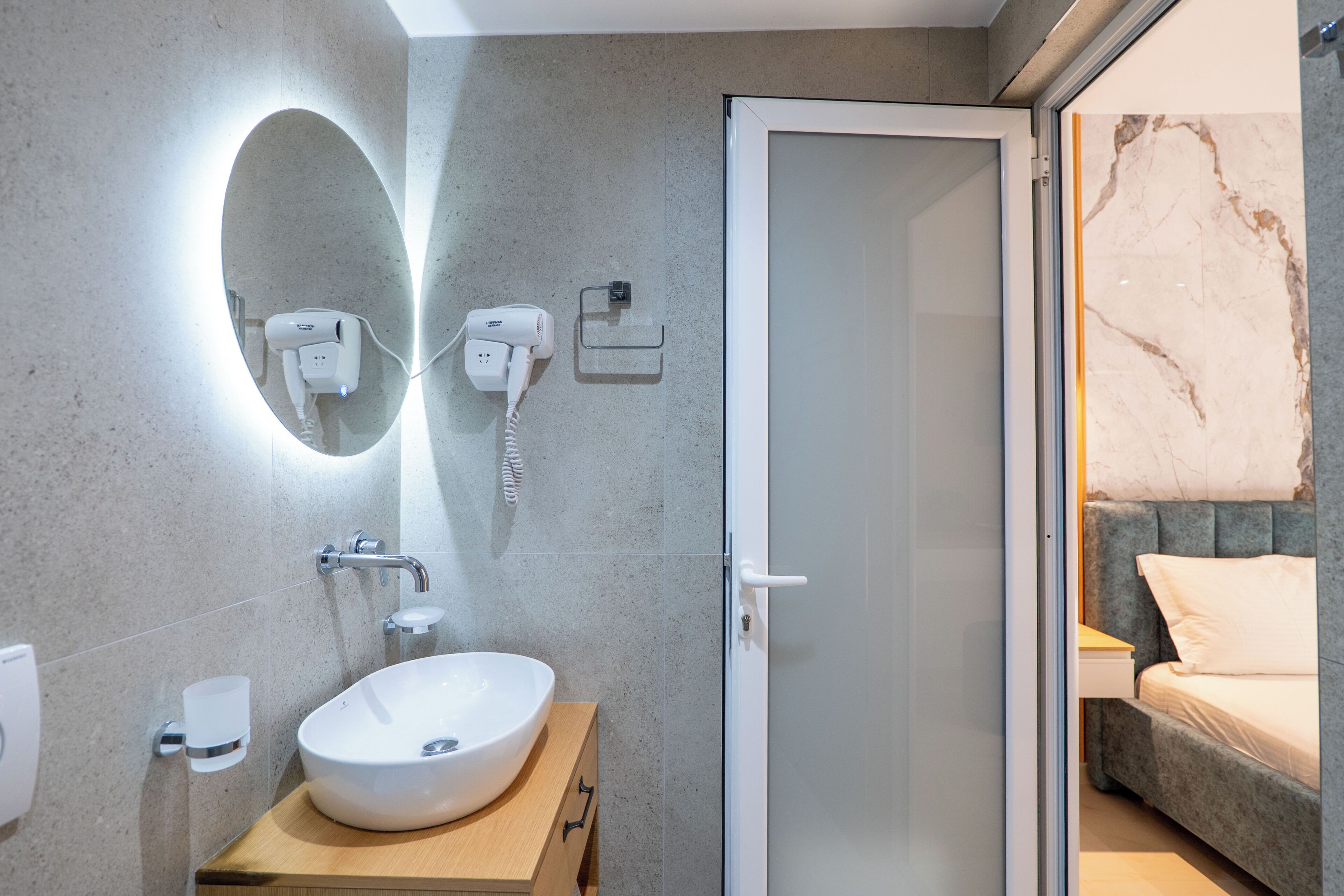 A modern bathroom with a large oval mirror, a white sink, and a handheld hair dryer on the wall.