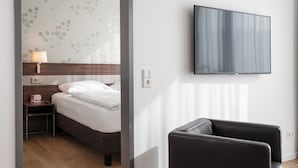 A hotel room with a bed, a nightstand with a lamp, a TV mounted on the wall, and a leather armchair.