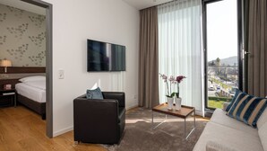 A modern hotel room with a bed, a sofa, a TV, a small table with flowers, and a view of the city through large windows.