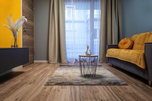 Ein modernes Wohnzimmer mit Holzboden, einem runden Couchtisch mit Drahtgestell, einem Sofa mit gelber Decke und einer Vase mit weißen Federn auf einem dunklen Konsolentisch.