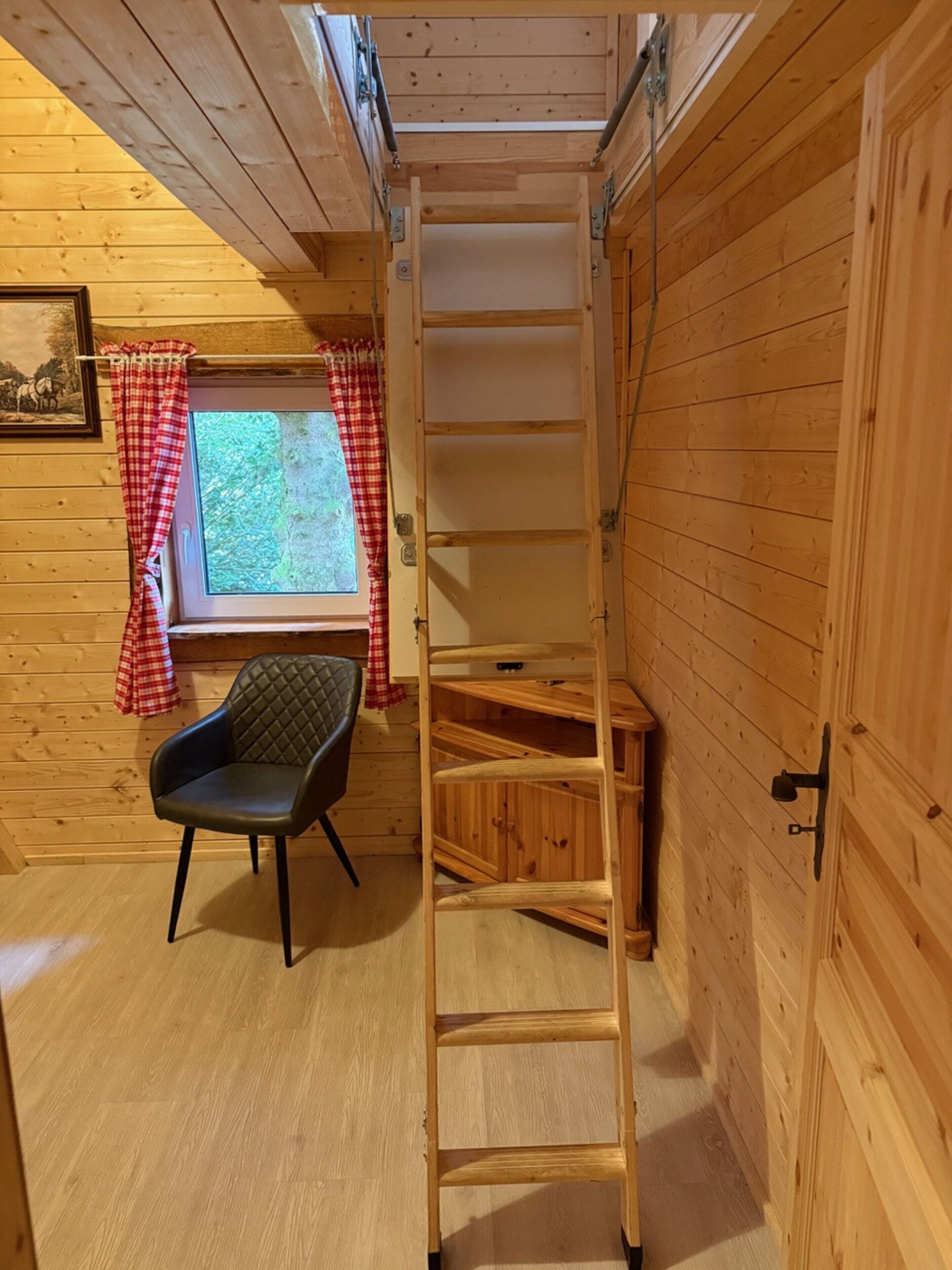 Una habitación tipo cabaña de madera con una cama en el altillo, una ventana con cortinas rojas, un sillón acolchado y una escalera de madera.