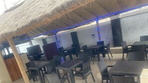Zona esterna con tavoli e sedie, tetto di paglia e illuminazione blu soffusa.