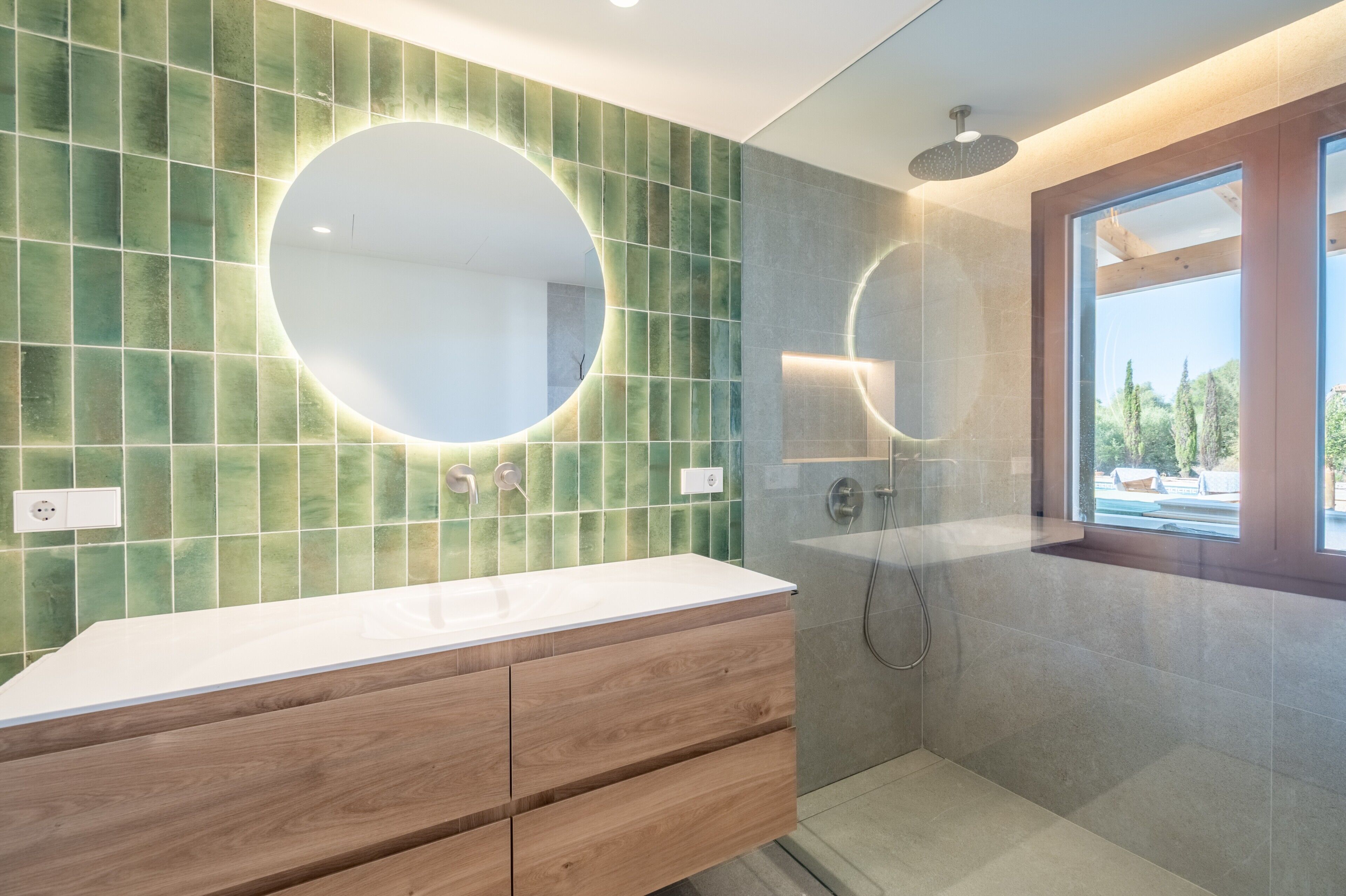 Une salle de bain moderne avec un mur recouvert de carreaux verts, un miroir rond, un plan de toilette blanc, des meubles en bois et un espace douche.