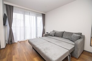 Un dormitorio moderno con un sofá cama gris, suelo de madera y un ventanal con cortinas translúcidas.