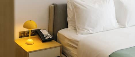 Un lit bien fait, avec du linge de lit blanc et des oreillers, une table de chevet avec une lampe et un téléphone, et une tête de lit grise.