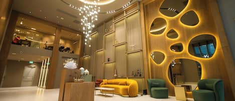 A modern lobby with a central yellow sofa, green armchairs, and a wooden reception desk.