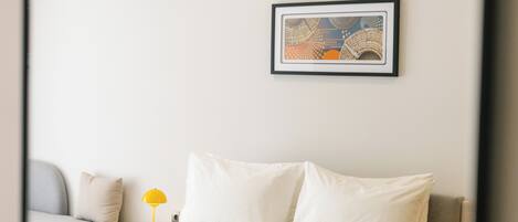 A neatly arranged bedroom with a bed, pillows, a bedside table with a lamp, and a framed artwork on the wall.