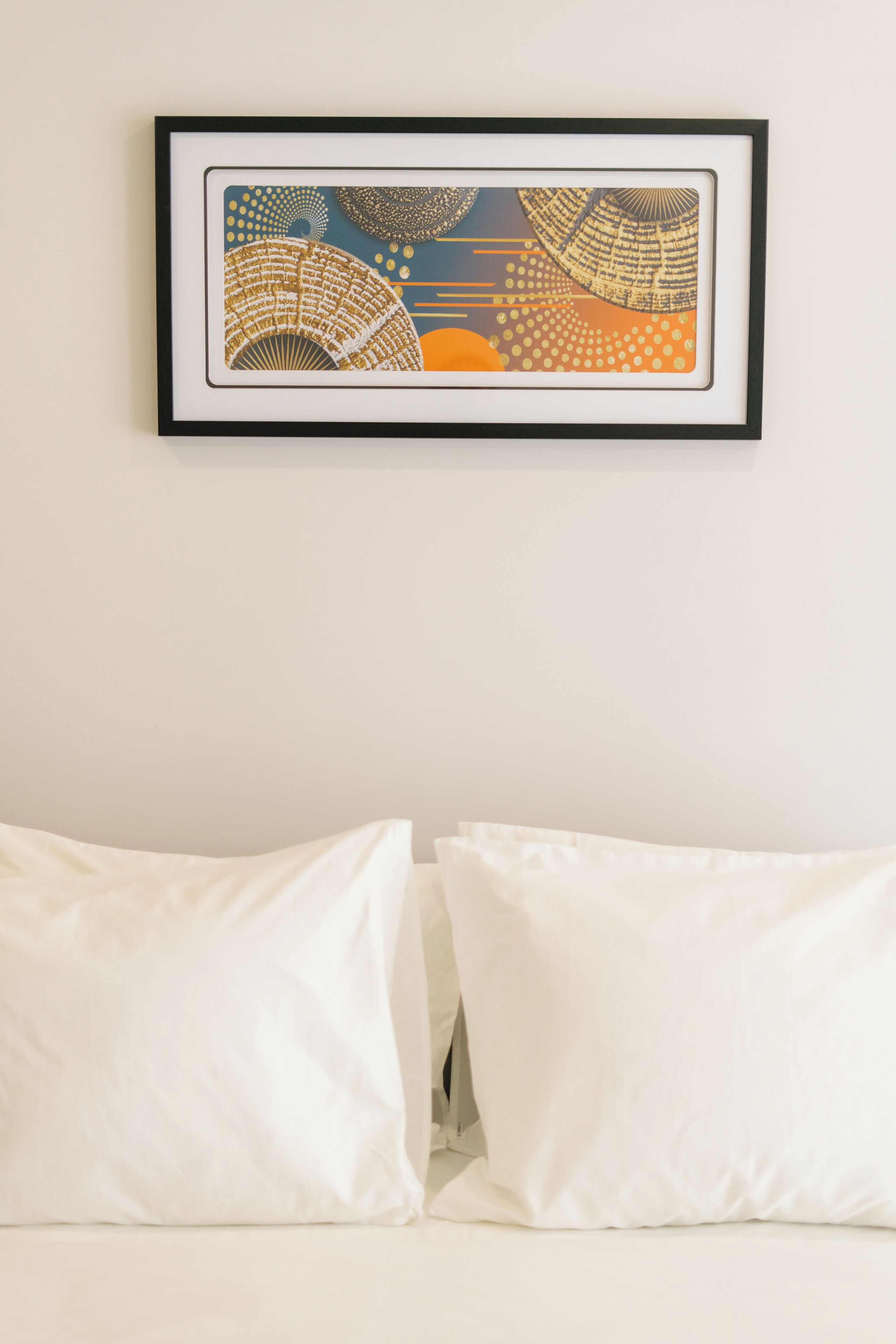 Une œuvre d’art abstraite encadrée est accrochée au mur au-dessus d’un lit où sont posés des oreillers blancs.