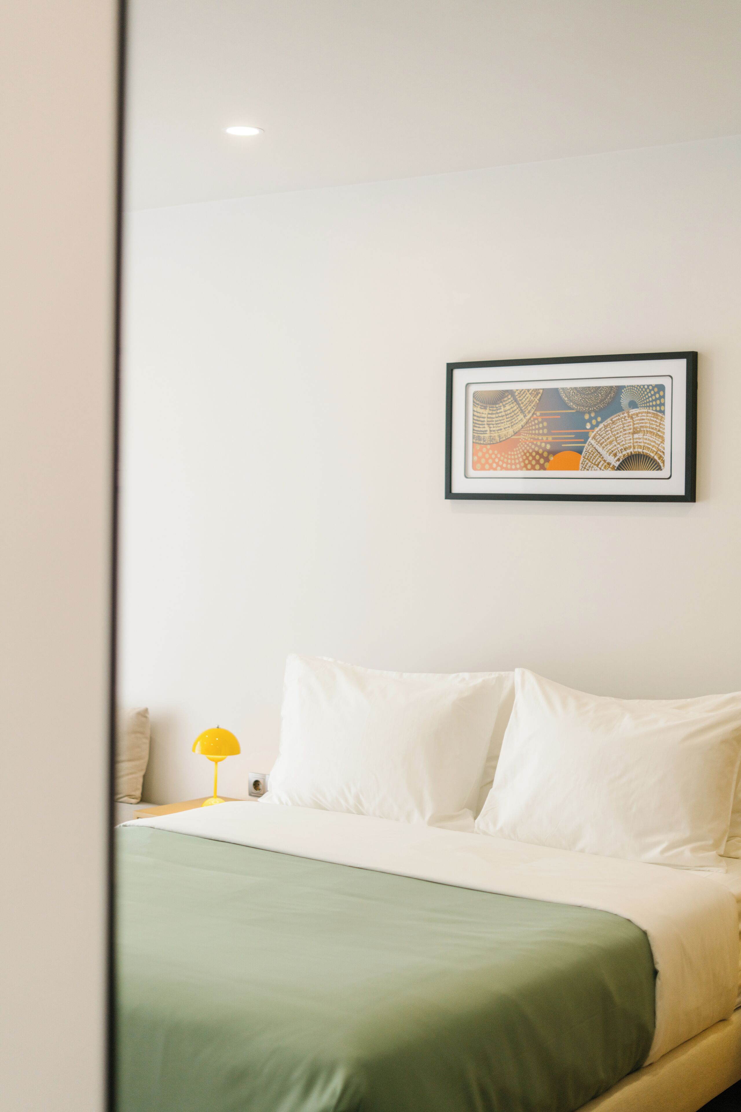 A neatly made bed with white pillows and a green bedspread, a framed abstract artwork on the wall, and a yellow bedside lamp.