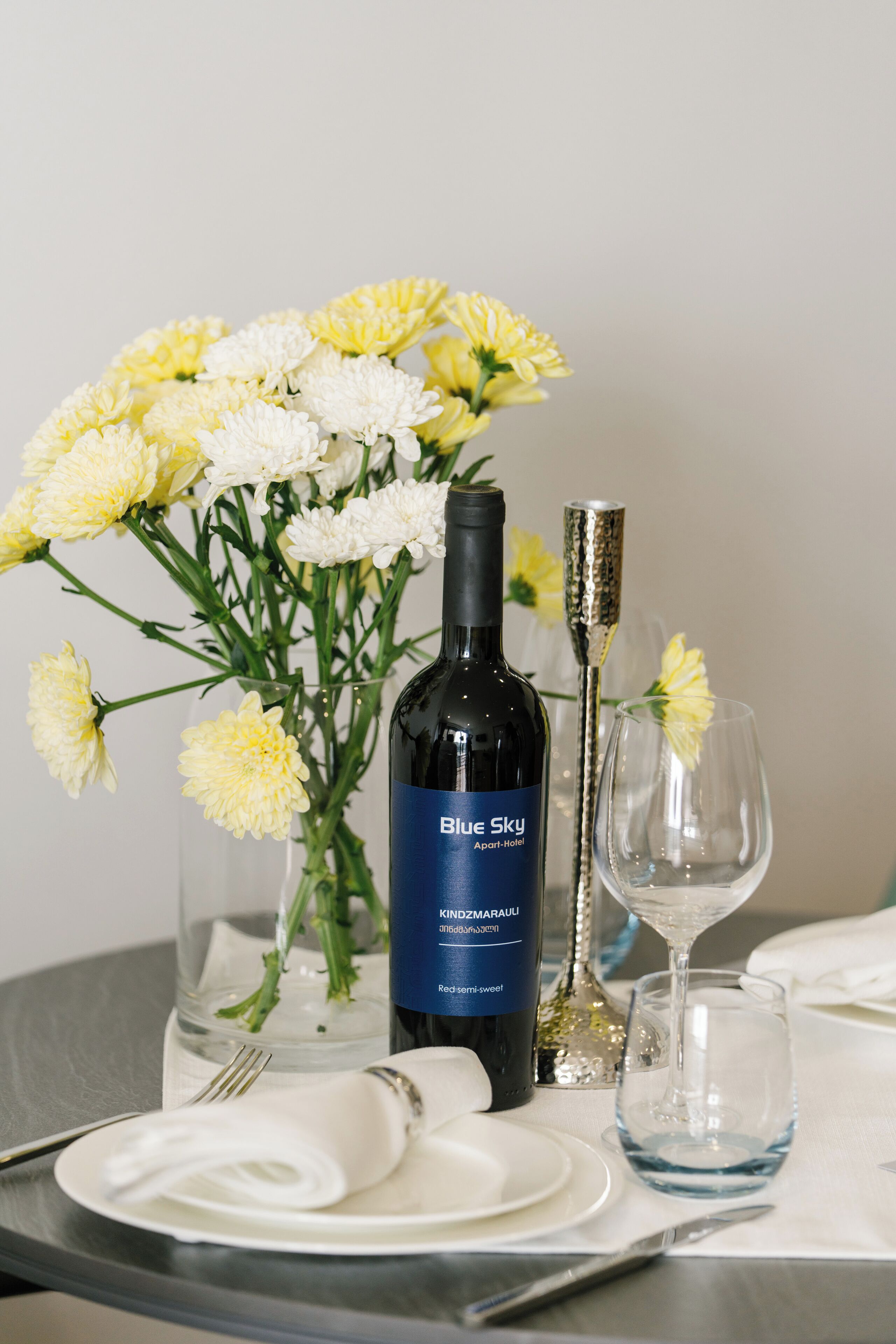 A table set with a bottle of wine, a glass, a vase with flowers, and a water glass.