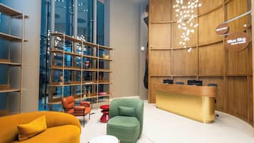 Un área de recepción moderna con un mostrador de madera, un sillón verde y un mostrador amarillo. Hay estantes con objetos decorativos y un taburete rojo.