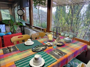 Un coin repas avec une table dressée pour deux personnes, une fenêtre donnant sur les arbres et une chaise avec un coussin.