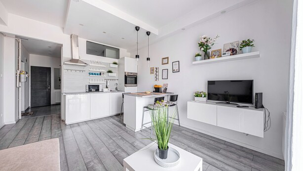 Une cuisine moderne avec des meubles blancs, un îlot central et un coin repas avec deux chaises. Un téléviseur à écran plat est fixé au mur, au-dessus d’une console blanche.