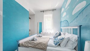 Une chambre à coucher avec un lit, une fenêtre ornée de rideaux légers et un mur sur lequel est accrochée une peinture abstraite bleue.