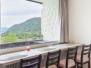 A room with a dining table, chairs, a window with a view of a forested hill, and a curtain. - Nice apartment for 3 people with A/C, WIFI, TV and balcony (Strobl)