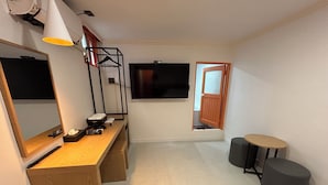 Ein kompakter Raum mit einem Holztisch, einem großen Spiegel, einem an der Wand befestigten Fernseher, einem kleinen Hocker und einer Tür zu einem weiteren Raum.