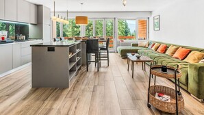 Een moderne keuken met een centraal eiland, een zithoek met een groene bank en uitzicht op groen via grote ramen.
