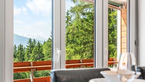 Een eetgedeelte met een tafel gedekt voor twee personen, grote ramen met uitzicht op een bos en bergen.