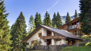 A modern chalet with a sloped roof, wooden balcony, and stone foundation, surrounded by forest and mountains.