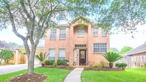 A two-story brick house with a front entrance, a covered porch, and a well-maintained lawn. - Modern 4BR Retreat Extended Stay Ideal. SW Houston (Sugar Land)