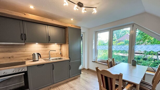 Een moderne keuken met grijze kasten, een houten aanrechtblad en een ingebouwde oven. Er is een eettafel met stoelen en uitzicht op groen buiten.