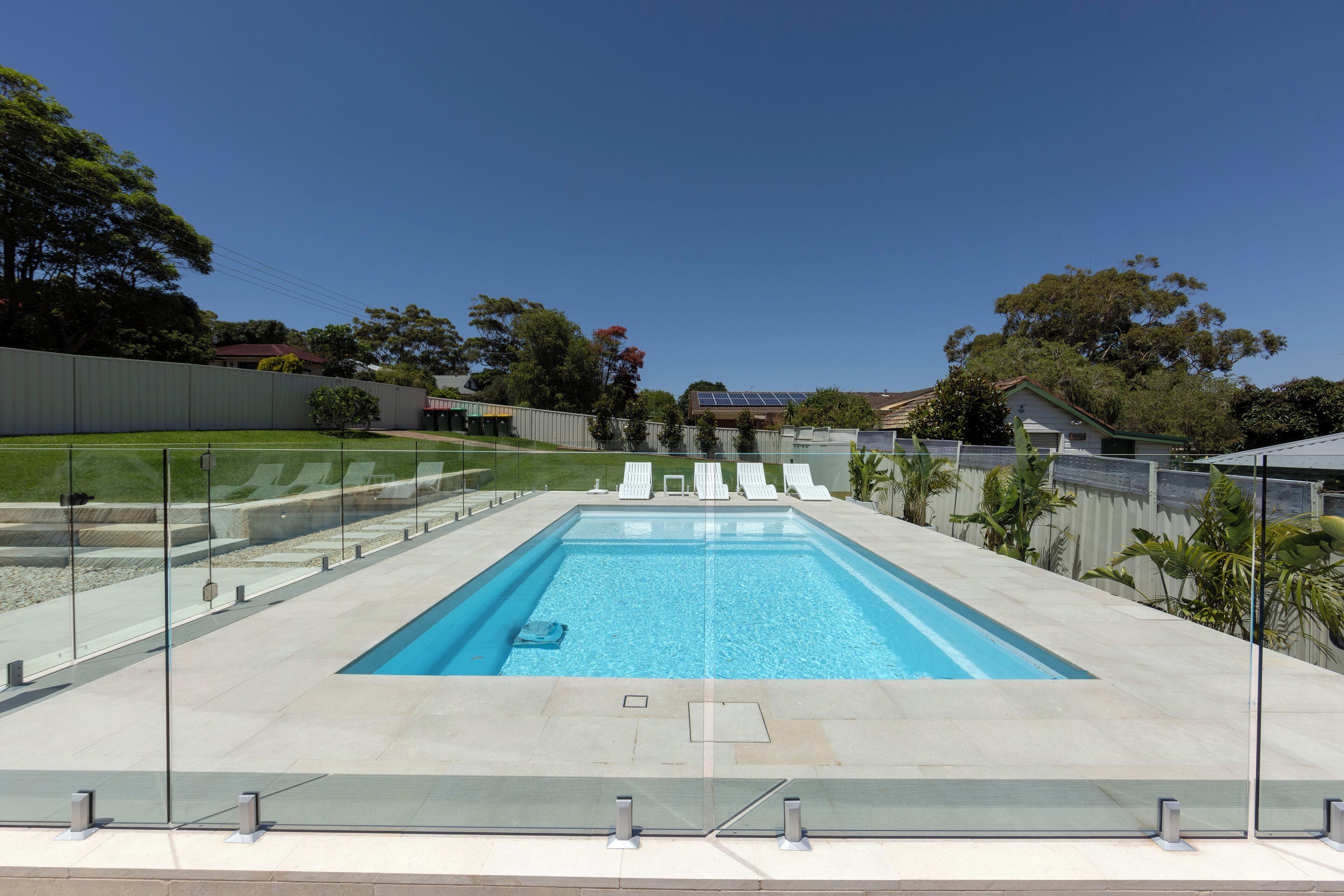 A rectangular swimming pool with clear blue water, surrounded by a glass fence, and a grassy area with trees and a fence in the background.