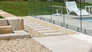 A modern outdoor pool area with a glass barrier, a white lounge chair, and a pebble-tiled walkway.