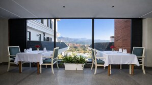 Un espace repas avec des tables et des chaises, une plante en pot, et une vue sur un paysage urbain et des montagnes, visible à travers de grandes fenêtres.