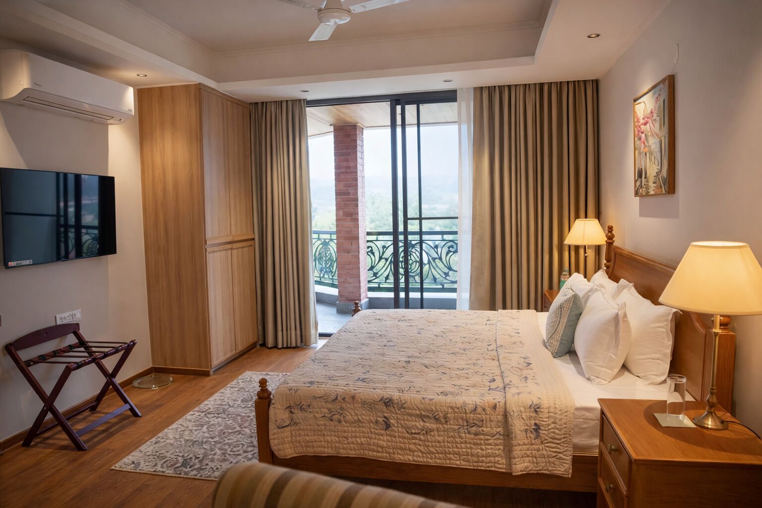 A hotel room with a large bed, a flat-screen TV, a wooden chair, a nightstand with a lamp, and a balcony with a view.