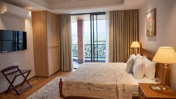 A hotel room with a large bed, a flat-screen TV, a wooden chair, a nightstand with a lamp, and a balcony with a view.