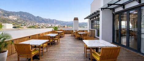 A rooftop terrace with wooden tables and chairs, a potted plant, and a view of a mountainous landscape.