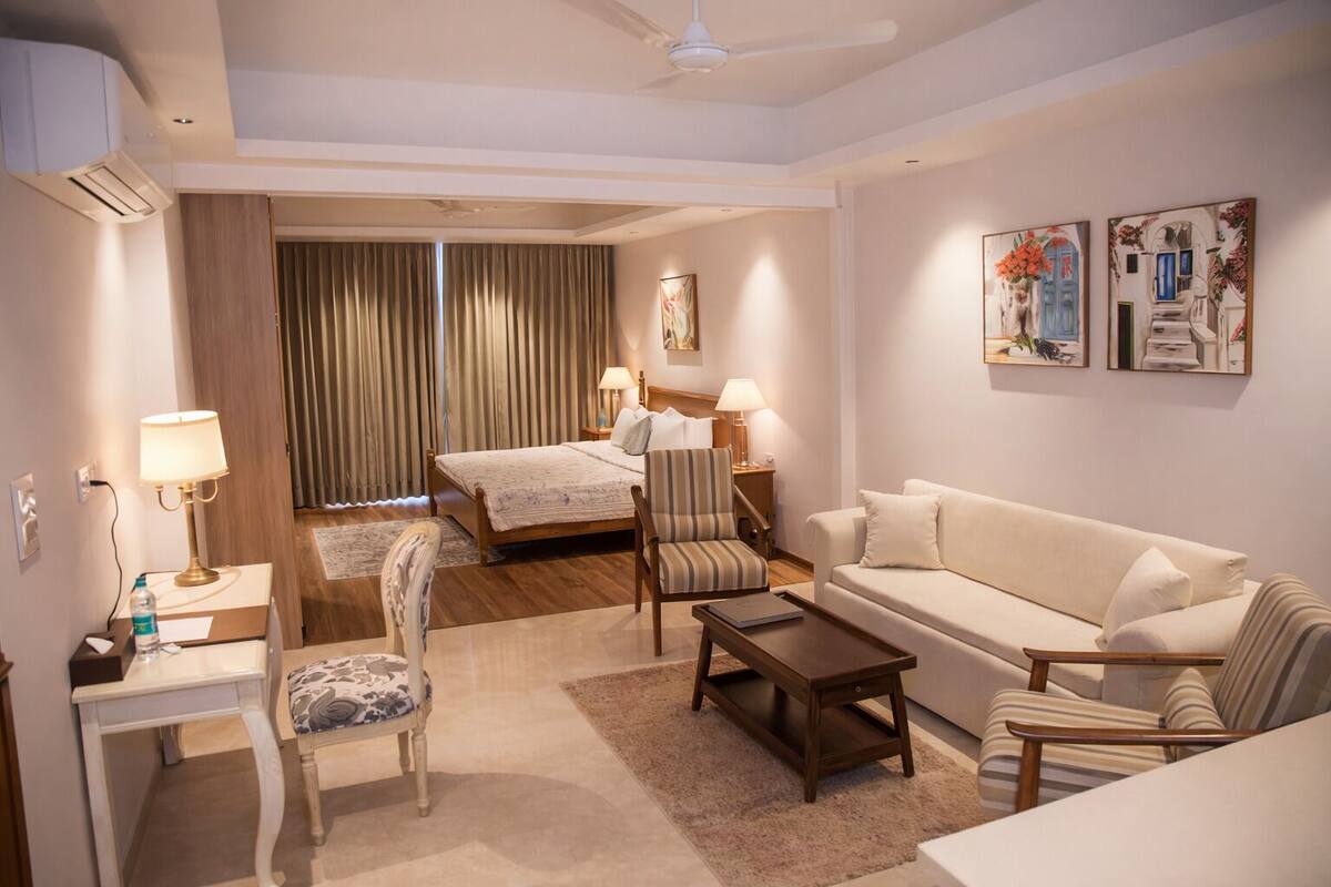 A hotel room with a bed, a sofa, a chair, a desk, and a coffee table. There are two lamps, a painting on the wall, and an air conditioning unit.