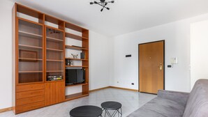 Una sala de estar con un sofá gris, dos taburetes negros, una estantería de madera y un televisor.