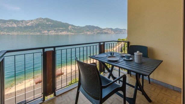 A balcony with a table and chairs overlooking a body of water and mountains. - Beautiful apartment for 4 guests with A/C, TV and patio (Torri Del Benaco)