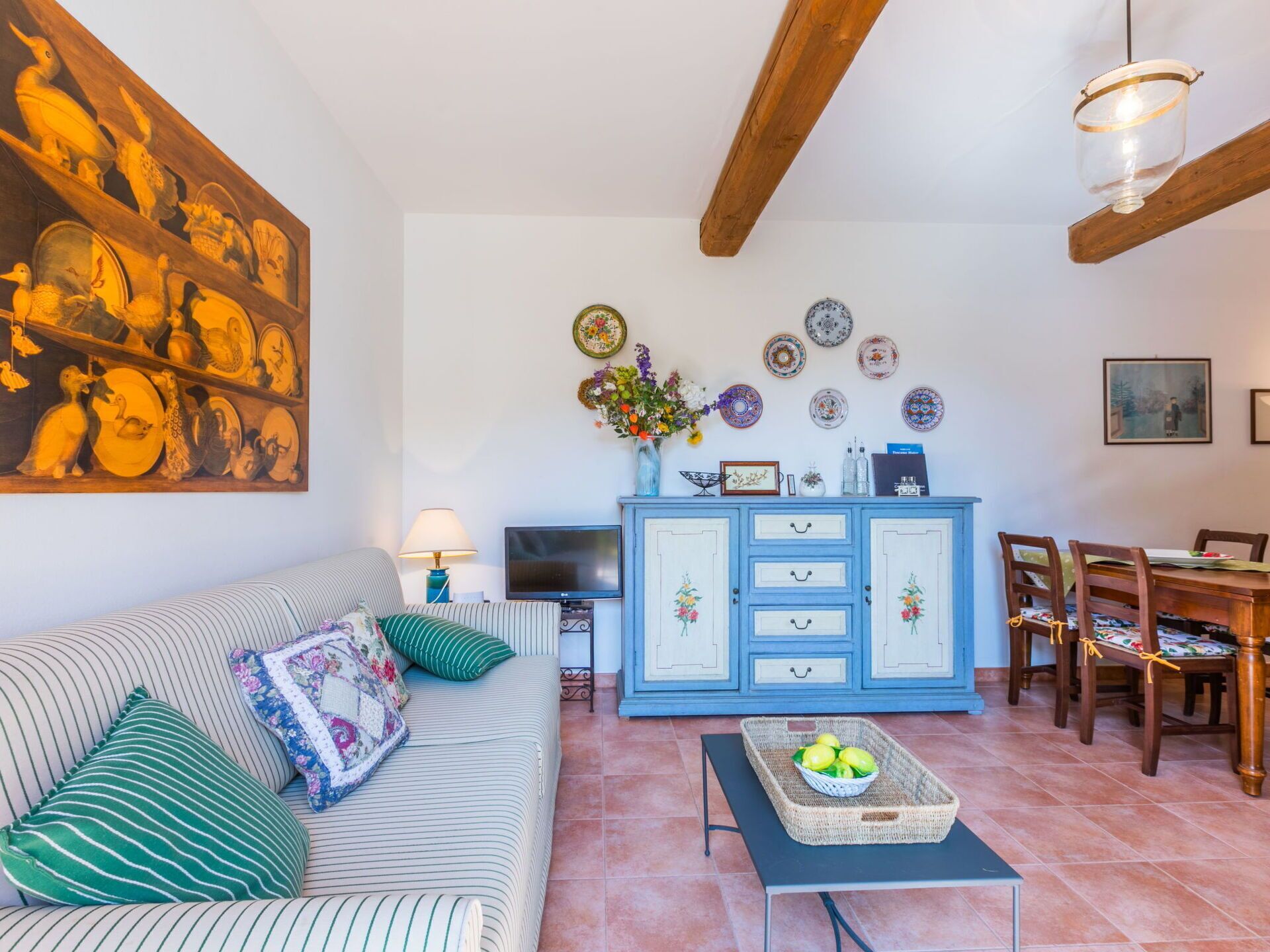 Un salon chaleureux avec un canapé à rayures, une table à manger entourée de chaises, un buffet orné d’assiettes décoratives et un tableau accroché au mur.