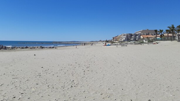 A wide sandy beach with a clear blue sky, a rocky breakwater, and buildings with balconies and palm trees in the background. - Large, comfortable T2 apartment (Mauguio)