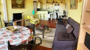 A living room with a sofa, a dining table with a floral tablecloth, a TV, and a kitchen area with cabinets and a dining table. - Large, comfortable T2 apartment (Mauguio)