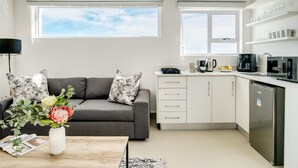 Un salon moderne avec un canapé gris, une table basse et un vase de fleurs. À côté se trouve une cuisine avec des meubles blancs, une machine à café noire et un four à micro-ondes.