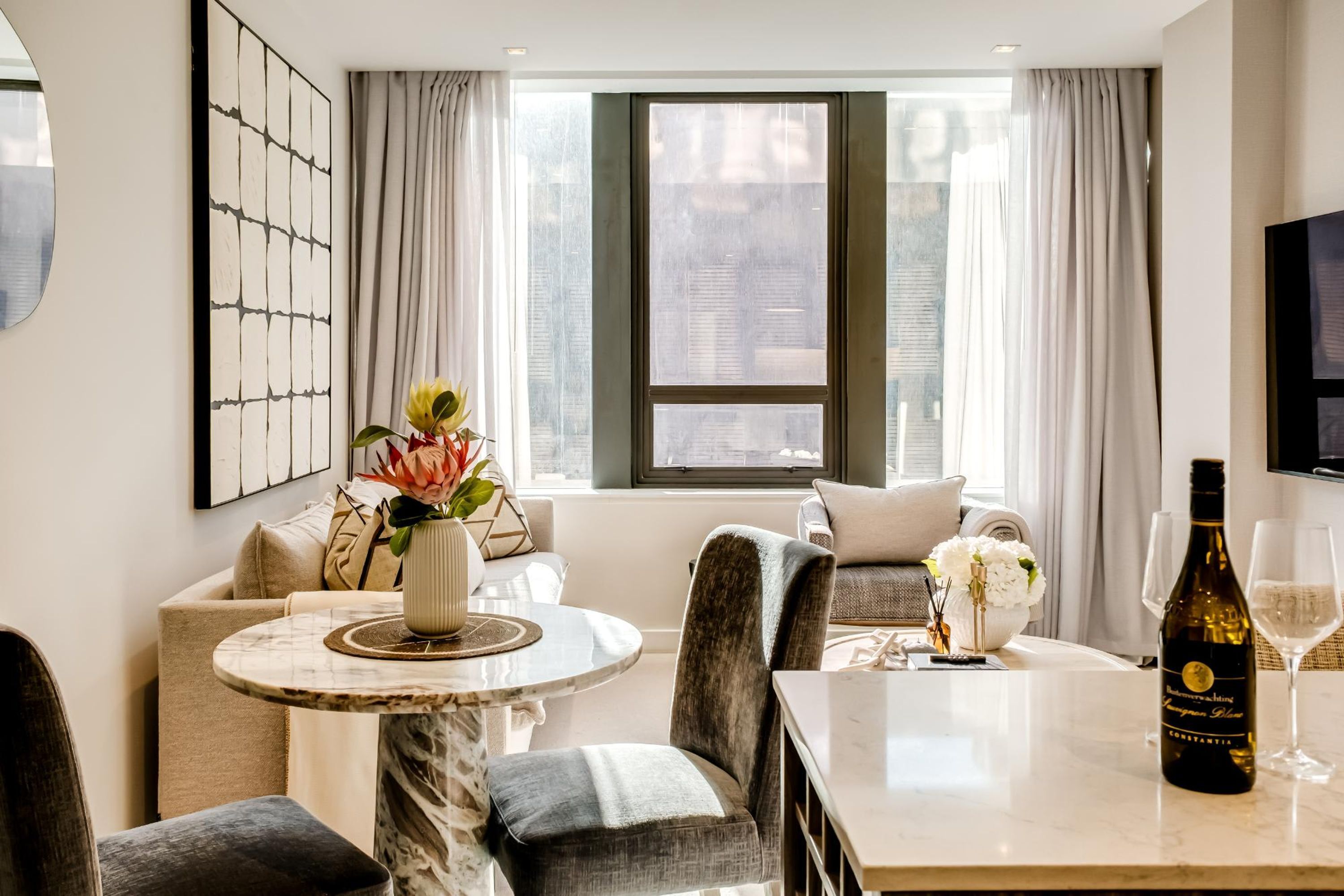 A modern hotel room with a dining area, a large window with curtains, a flat-screen TV, and a bottle of wine on the table.