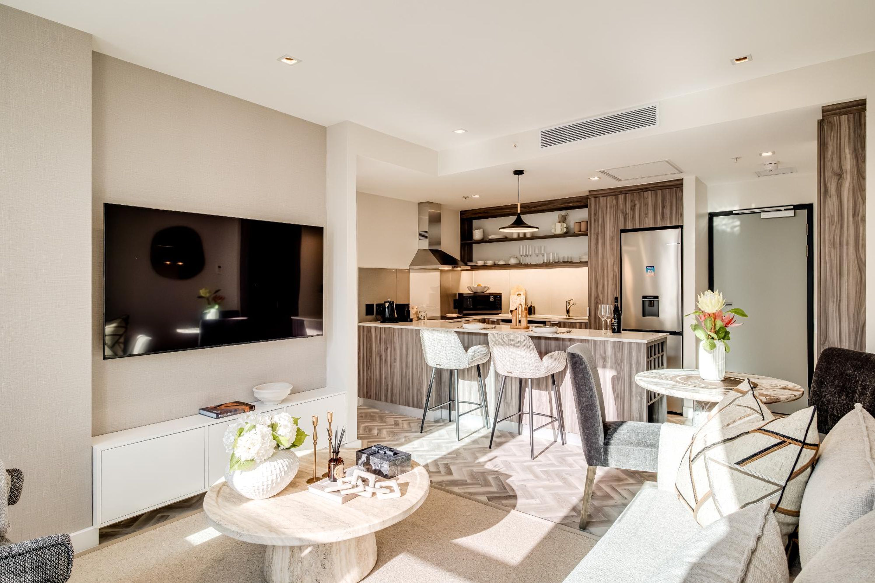 Una sala de estar moderna con espacio para la cocina, que incluye una mesa de comedor, taburetes de bar y un televisor de pantalla plana montado en la pared.