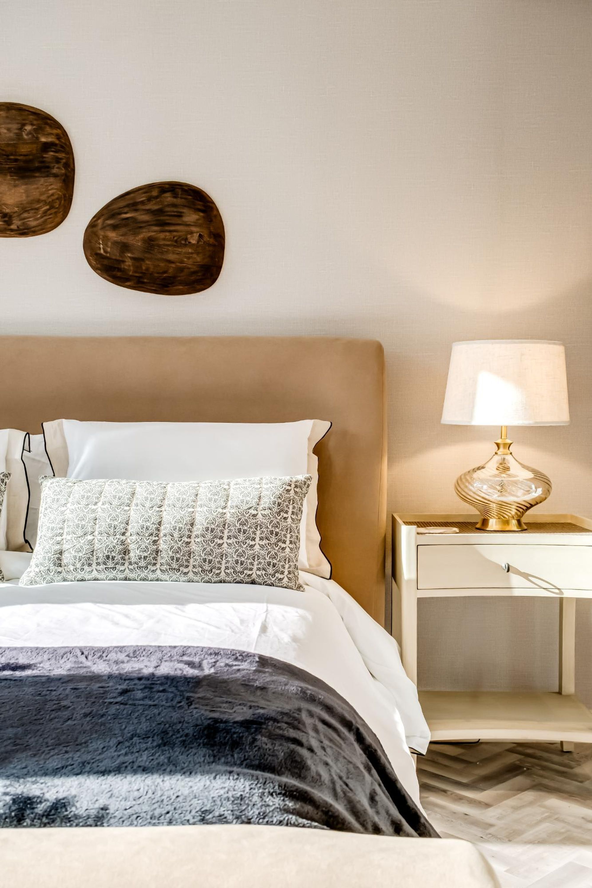 A neatly made bed with a white and gray patterned pillow, a dark throw blanket, and a gold-rimmed lamp on a bedside table.
