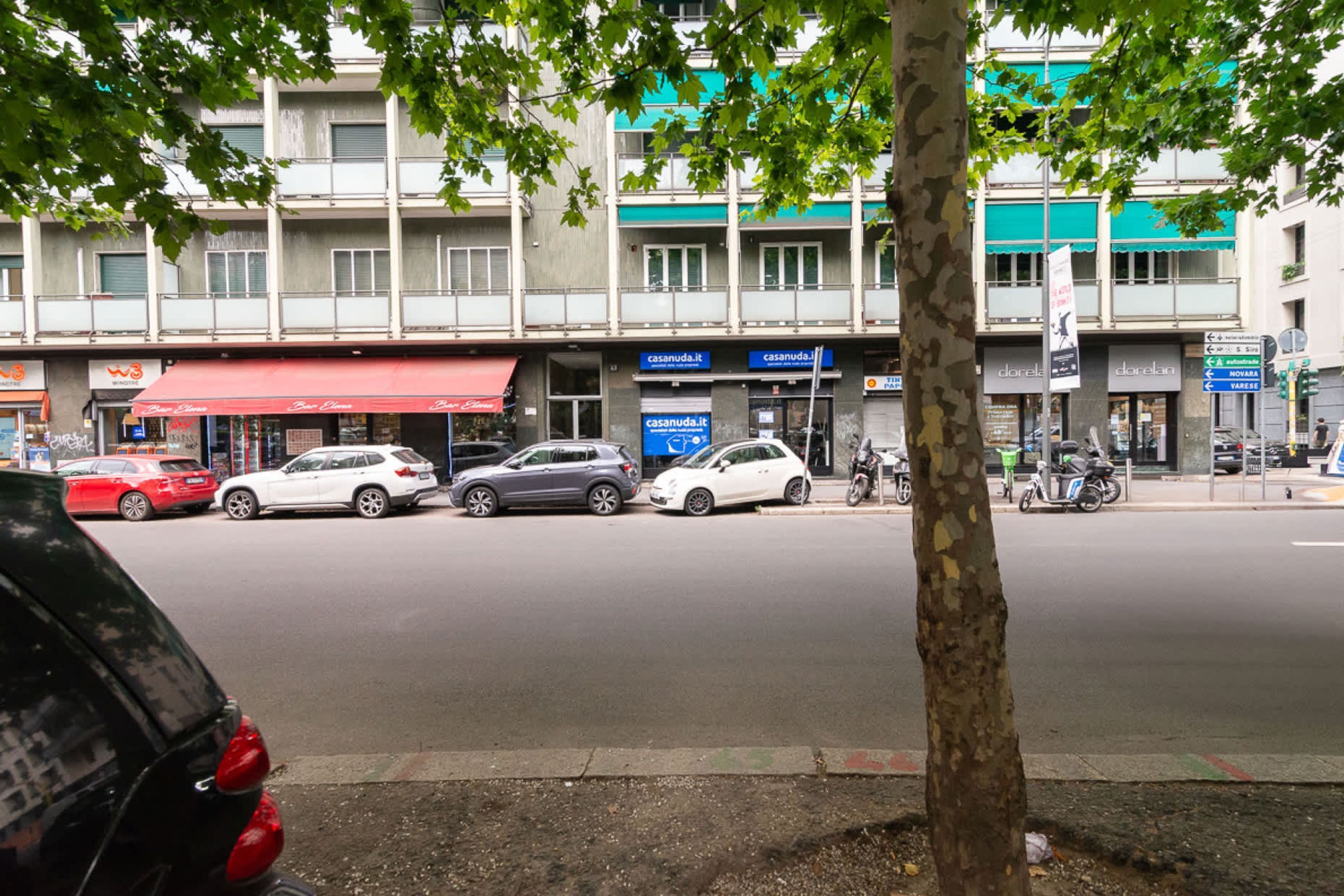 Straßenansicht mit parkenden Autos, einem Baum und Gebäuden mit Schaufenstern.