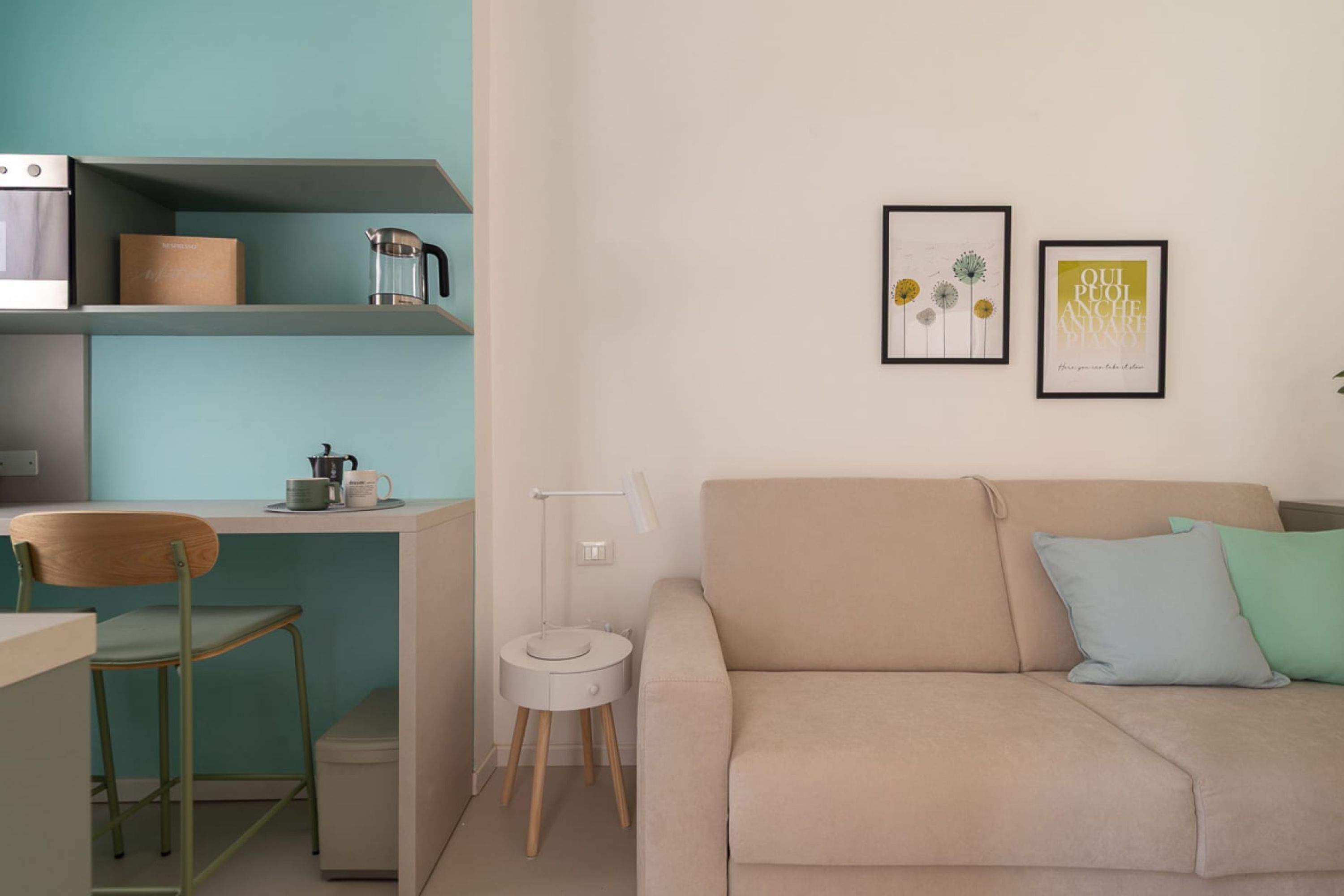 Un salon moderne avec un canapé beige, une chaise en bois, une petite table avec une bouilloire et une lampe fixée au mur.