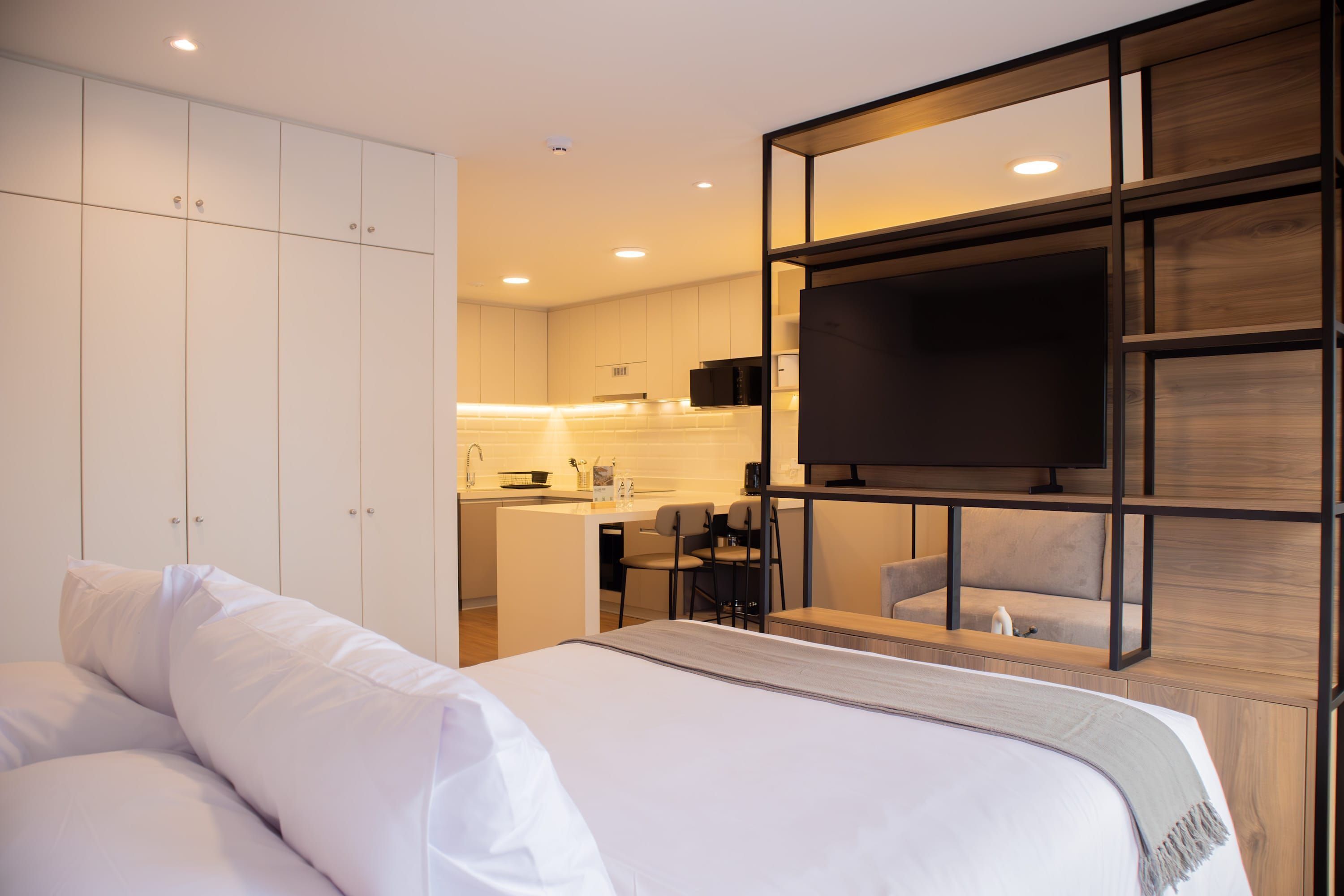 A modern hotel room with a large bed, a TV, a kitchenette, and a seating area.