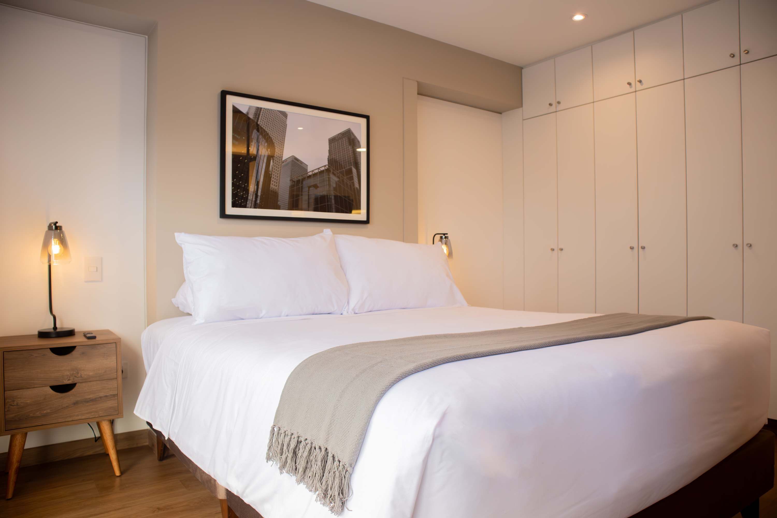 A neatly made bed with white linens and a grey throw blanket, a wooden nightstand with a lamp, and a framed picture on the wall.